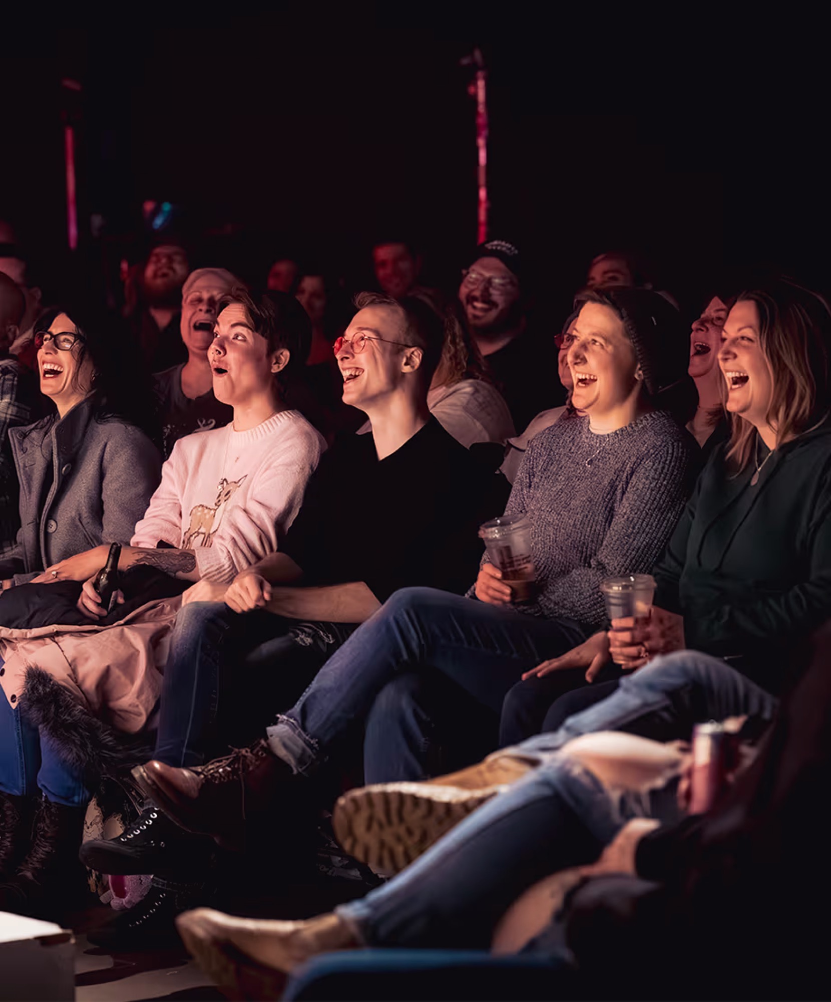 A packed front row mid-laugh, drinks in hand, during a roast battle.
