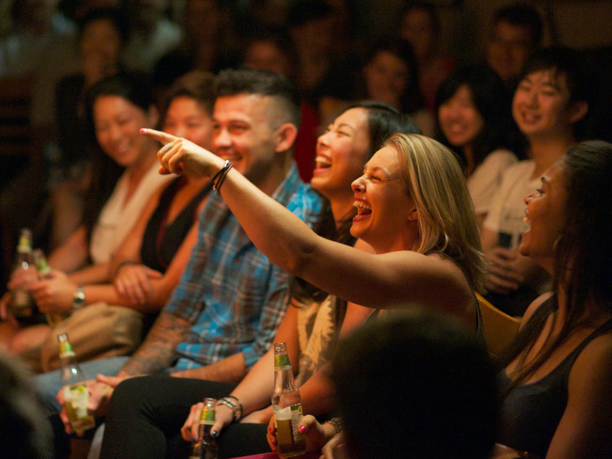 A packed front row mid-laugh, drinks in hand, lit warm from the stage.
